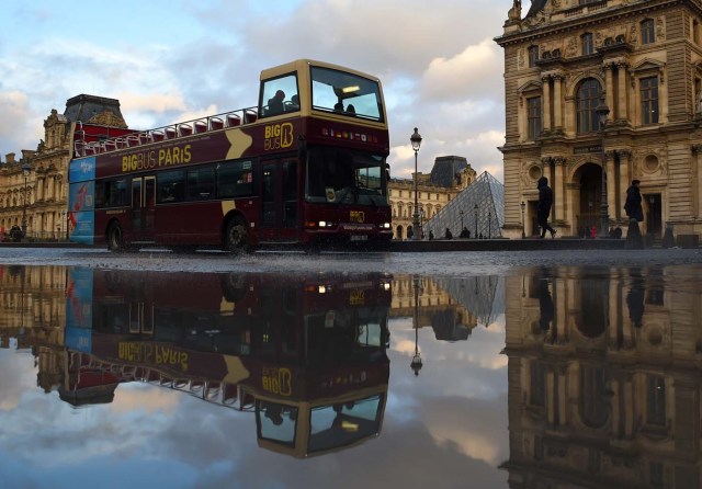 A double decker sightseeing bus drives past the Louvre Museum, reflected in a pool of rain water on January 1, 2018 in Paris. / AFP PHOTO / GUILLAUME SOUVANT